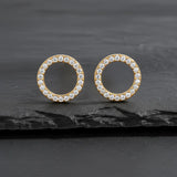 Runde vergoldete Damen Ohrstecker in offener Kreisform, komplett mit klar funkelnden Zirkonia besetzt, elegant inszeniert auf dunklem Schieferhintergrund.