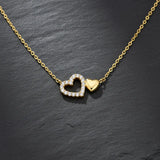 Vergoldete Damen Halskette mit zwei verbundenen Herzanhängern: ein pavé-besetztes Zirkonia-Herz und ein poliertes Goldherz, elegant präsentiert auf schwarzem Schieferhintergrund.