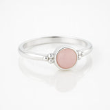 Silberring mit rundem rosafarbenem Stein Rosenquarz und kleinen Silberkügelchen neben der Fassung, liegend auf einem glänzenden weißen Untergrund mit weicher Spiegelung.