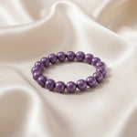 Ein Armbands aus glänzenden, lila Perlen, das auf einem beigefarbenen, seidenartigen Stoff liegt.