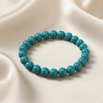 Ein Armbands aus glänzenden, türkisfarbenen Perlen, das auf einem beigefarbenen, seidenartigen Stoff liegt.