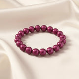 Ein Armbands aus glänzenden, pflaumenfarbenen Perlen, das auf einem beigefarbenen, seidenartigen Stoff liegt.