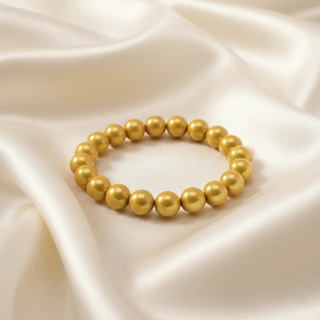 Ein Armbands aus glänzenden, goldfarbenen Perlen, das auf einem beigefarbenen, seidenartigen Stoff liegt.