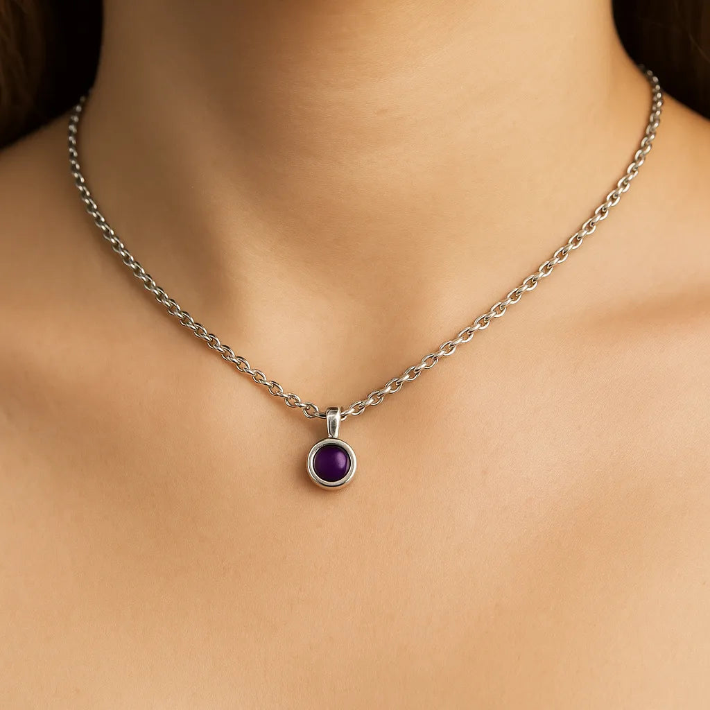 Nahaufnahme eines Frauenhalses mit einer silbernen Kette und einem kleinen, runden Amethyst-Cabochon-Anhänger von 5 mm Durchmesser. Der Anhänger zeigt einen tiefvioletten, glatten Stein in einer polierten Silberfassung. Der Hintergrund ist neutral und weich ausgeleuchtet, der Fokus liegt klar auf dem Anhänger.