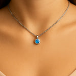 Nahaufnahme eines Frauenhalses mit einer silbernen Kette und einem kleinen, runden blauen Calcedon-Cabochon-Anhänger von 5 mm Durchmesser. Der sanft schimmernde, himmelblaue Stein ist in einer glänzenden Silberfassung eingefasst. Der Hintergrund ist neutral und weich ausgeleuchtet, der Fokus liegt auf dem Anhänger.