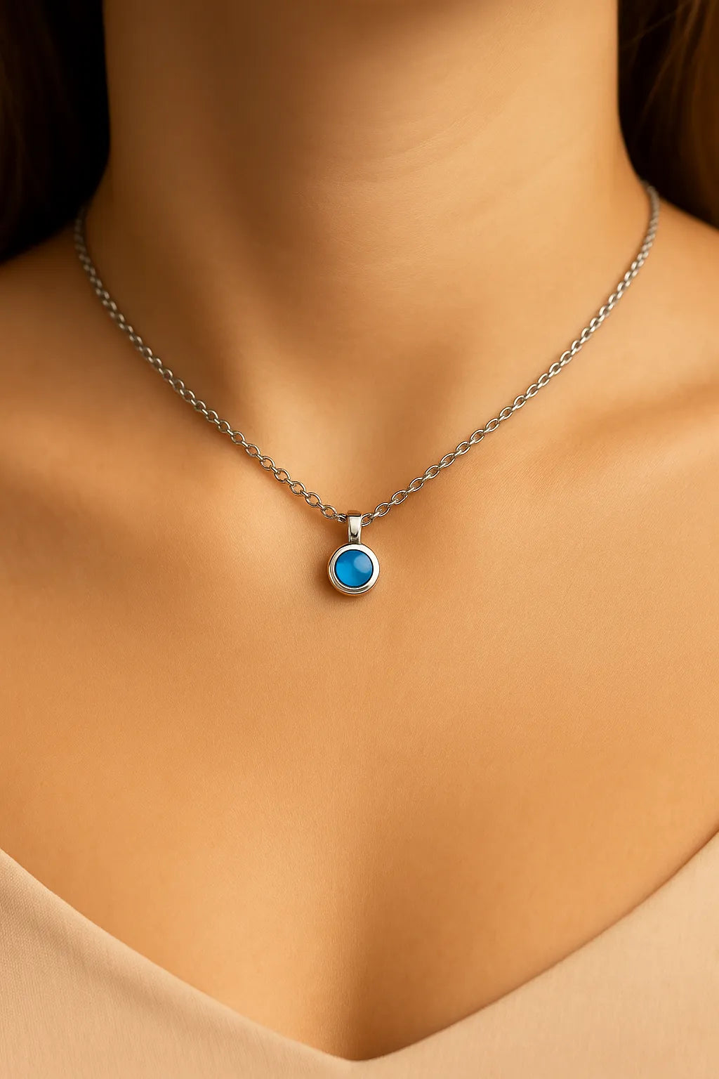 Nahaufnahme eines Frauenhalses mit einer silbernen Kette und einem kleinen, runden blauen Calcedon-Cabochon-Anhänger von 5 mm Durchmesser. Der sanft schimmernde, himmelblaue Stein ist in einer glänzenden Silberfassung eingefasst. Der Hintergrund ist neutral und weich ausgeleuchtet, der Fokus liegt auf dem Anhänger.