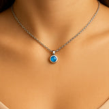 Nahaufnahme eines Frauenhalses mit einer silbernen Kette und einem kleinen, runden blauen Calcedon-Cabochon-Anhänger von 5 mm Durchmesser. Der sanft schimmernde, himmelblaue Stein ist in einer glänzenden Silberfassung eingefasst. Der Hintergrund ist neutral und weich ausgeleuchtet, der Fokus liegt auf dem Anhänger.