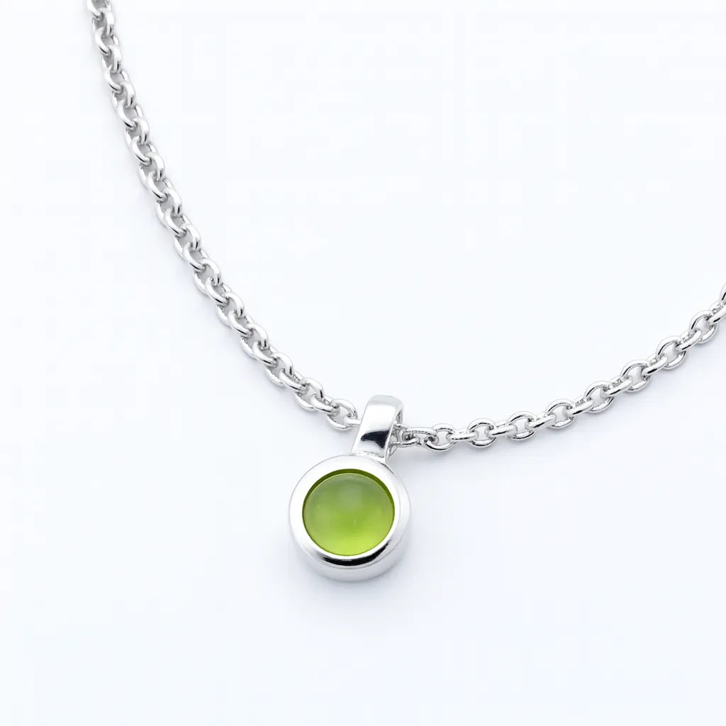 Nahaufnahme einer silbernen Halskette mit einem kleinen, runden Peridot-Cabochon-Anhänger. Der hellgrüne, glatte Edelstein ist in einer polierten Silberfassung eingefasst und hängt an einer feinen Gliederkette auf weißem Hintergrund. Der Fokus liegt auf dem Anhänger, der schlicht und elegant wirkt.
