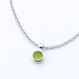 Nahaufnahme einer silbernen Halskette mit einem kleinen, runden Peridot-Cabochon-Anhänger. Der hellgrüne, glatte Edelstein ist in einer polierten Silberfassung eingefasst und hängt an einer feinen Gliederkette auf weißem Hintergrund. Der Fokus liegt auf dem Anhänger, der schlicht und elegant wirkt.