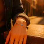 Eine Frau trägt ein silbernes Armband mit fünf farbigen Edelsteinen an ihrem Handgelenk. Ihr Arm ruht auf einem warm beleuchteten Stein, im Hintergrund eine sonnige urbane Szenerie mit sanft verschwommenen Gebäuden.