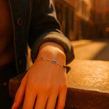 Eine Frau trägt ein silbernes Armband mit fünf farbigen Edelsteinen an ihrem Handgelenk. Ihr Arm ruht auf einem warm beleuchteten Stein, im Hintergrund eine sonnige urbane Szenerie mit sanft verschwommenen Gebäuden.