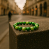 Ein Armband aus glänzenden hell- und dunkelgrünen Perlen liegt auf einem steinernen Podest in einer Stadtstraße. Der Hintergrund mit Gebäuden und Lichtern ist unscharf, wodurch das Armband im Vordergrund betont wird.