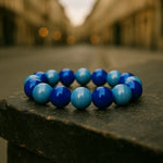 Ein Armband aus glänzenden blauen und hellblauen Perlen liegt auf einem steinernen Podest in einer Stadtstraße. Der Hintergrund mit Gebäuden und warmen Lichtern ist unscharf, wodurch das Armband im Vordergrund hervorgehoben wird.