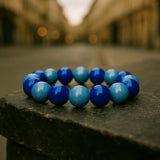 Ein Armband aus glänzenden blauen und hellblauen Perlen liegt auf einem steinernen Podest in einer Stadtstraße. Der Hintergrund mit Gebäuden und warmen Lichtern ist unscharf, wodurch das Armband im Vordergrund hervorgehoben wird.