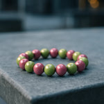 Ein glänzendes Armband aus abwechselnd rosa und grünen Perlen liegt auf einer kühlen, grauen Betonoberfläche in urbaner Umgebung. Der Hintergrund ist weich verschwommen, wodurch das Armband klar im Fokus steht. Das diffuse Licht betont den Perlglanz und schafft eine ruhige, moderne Atmosphäre.