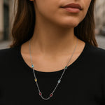 Eine junge Frau steht in einer urbanen Umgebung und trägt eine zarte Silberkette mit rechteckigen Edelsteinen in den Farben Hellblau, Grün, Gelb, Hellblau und zwei Rottönen. Die Kette liegt elegant auf einem schwarzen Oberteil und wird durch das natürliche Licht dezent hervorgehoben. Der Hintergrund ist leicht unscharf, wodurch der Fokus klar auf der Halskette liegt.