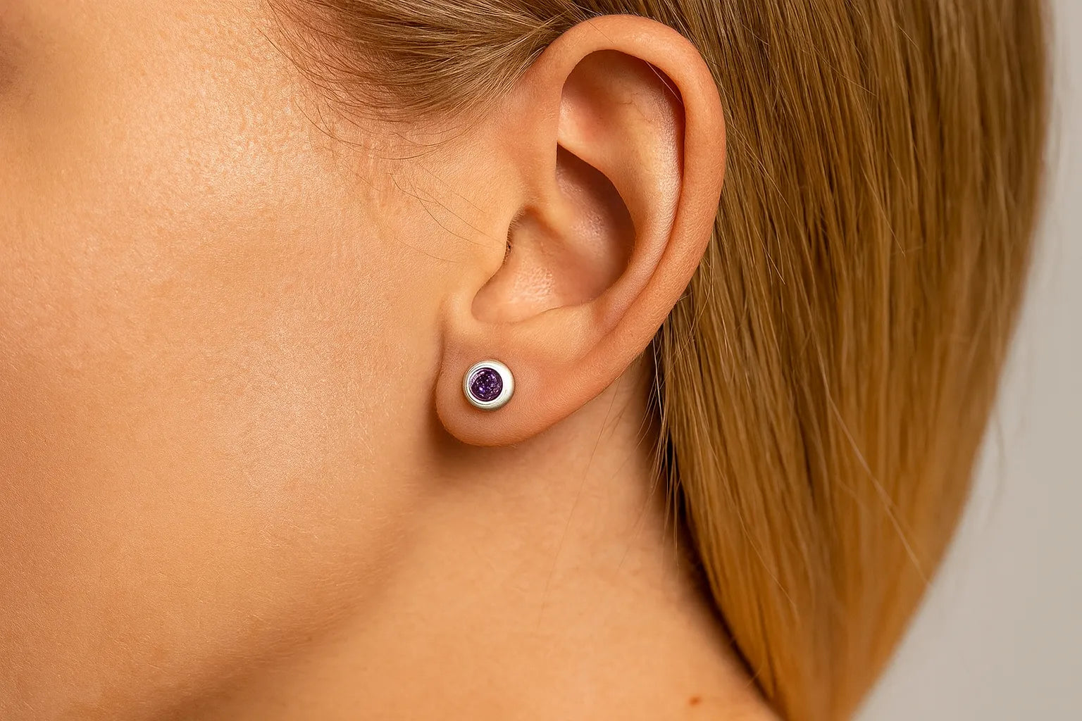 Nahaufnahme einer Frau im Profil mit einem kleinen runden Ohrstecker aus Silber und einem violetten Amethyst im Ohr. Der facettierte Edelstein funkelt im Licht, während die glatte, helle Haut und das zurückgelegte blonde Haar den Ohrring deutlich hervorheben