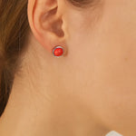 Nahaufnahme einer Frau in Seitenansicht mit einem kleinen silbernen Ohrstecker, der eine glänzende, korallenrote Kuppel zeigt. Der Ohrring hat einen Durchmesser von etwa 5 mm und sitzt dezent am Ohrläppchen, während Haar und Haut in warmem Licht natürlich wirken.