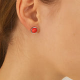 Nahaufnahme einer Frau in Seitenansicht mit einem kleinen silbernen Ohrstecker, der eine glänzende, korallenrote Kuppel zeigt. Der Ohrring hat einen Durchmesser von etwa 5 mm und sitzt dezent am Ohrläppchen, während Haar und Haut in warmem Licht natürlich wirken.