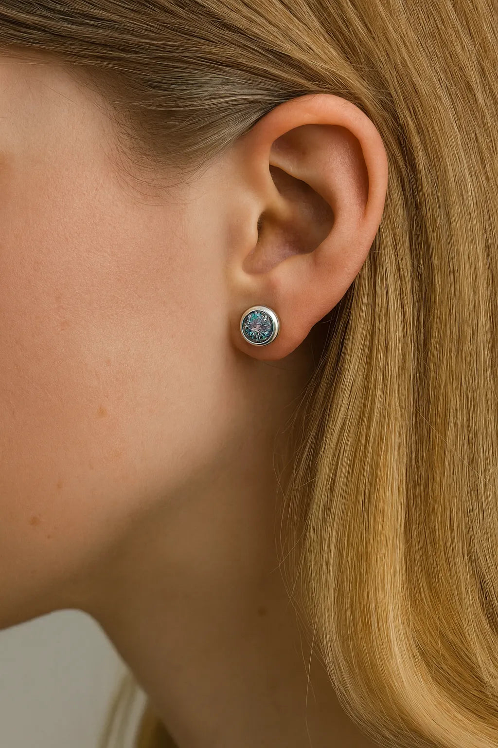 Nahaufnahme einer Frau im Profil mit einem kleinen runden Ohrstecker aus Silber und einem facettierten mystischen Topas im Ohr. Der Stein zeigt schillernde Farbreflexe in Lila-, Blau- und Grüntönen, die im Licht glitzern. Das blonde Haar ist hinter das Ohr gelegt, um den funkelnden Ohrring deutlich hervorzuheben.