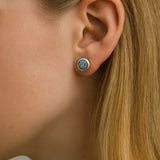 Nahaufnahme einer Frau im Profil mit einem kleinen runden Ohrstecker aus Silber und einem facettierten mystischen Topas im Ohr. Der Stein zeigt schillernde Farbreflexe in Lila-, Blau- und Grüntönen, die im Licht glitzern. Das blonde Haar ist hinter das Ohr gelegt, um den funkelnden Ohrring deutlich hervorzuheben.