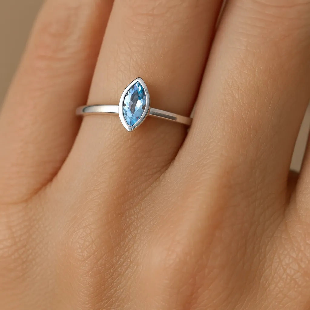 Ein eleganter Damenring aus poliertem 925er Sterlingsilber mit einem 5 mm langen, facettierten blauen Topas im Navette-Schliff. Der Ring ist an der Hand einer Frau am Ringfinger zu sehen. Der Fokus liegt auf dem hellblau funkelnden Edelstein, der von einer schlichten, glänzenden Ringschiene gehalten wird.