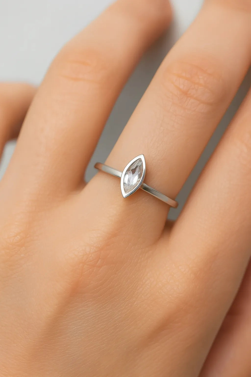 Eine Nahaufnahme einer Frauenhand, die einen eleganten Silberring mit einem kleinen, länglich facettierten Mondstein am Ringfinger trägt. Der transparente Stein zeigt zarte Blauschimmer, eingefasst in eine schlichte, glänzende Fassung. Der Fokus liegt klar auf dem Ring, während der Hintergrund neutral und weich verschwommen ist.