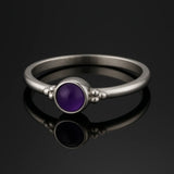 Sterlingsilber-Ring mit poliertem Amethyst-Cabochon in sattem Violett, fotografiert auf dunklem glänzenden Untergrund mit klarer Spiegelung für hochwertige Schmuckpräsentation.