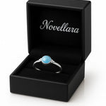 Eleganter Damenring aus Sterling Silber mit rundem blauem Chalcedon Cabochon (4 mm) in Zargenfassung, präsentiert in geöffneter schwarzer Schmuckbox mit Novellara Logo – hochwertiger Silberring mit hellblauem Edelstein als Geschenkidee für Frauen.