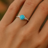 Eine Nahaufnahme einer Frauenhand zeigt einen eleganten Silberring mit einem runden, himmelblauen Chalcedon-Cabochon. Der 4 mm große Stein ist glatt poliert und in eine schlichte Silberfassung mit kleinen Kugeldetails eingefasst. Natürliches Licht hebt den sanften Glanz des Steins und der Ringschiene hervor, während der neutrale Hintergrund den Fokus auf den Ring lenkt.