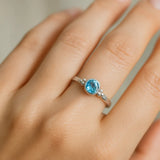 Eine Nahaufnahme einer Frauenhand zeigt einen eleganten Silberring mit einem runden, facettierten Blautopas. Der hellblaue Edelstein mit 4 mm Durchmesser ist in eine glatte Fassung eingefasst und wird von kleinen silbernen Kugeldetails flankiert. Natürliches, weiches Licht betont den klaren Glanz des Steins und die polierte Oberfläche der Ringschiene vor einem neutralen, unscharfen Hintergrund.