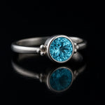 Sterlingsilber-Ring mit brillant geschliffenem blauem Topas, seitliche Ansicht auf schwarzem kontrastreichen Spiegel-Untergrund, professionelle Schmuckfotografie für Produktdarstellung.