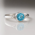 Feiner Silberring mit facettiertem Swiss Blue Topas, liegend auf hellem glänzendem Untergrund mit Spiegelung, hochwertige Nahaufnahme von handgefertigtem Damenschmuck.