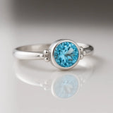 Feiner Silberring mit facettiertem Swiss Blue Topas, liegend auf hellem glänzendem Untergrund mit Spiegelung, hochwertige Nahaufnahme von handgefertigtem Damenschmuck.