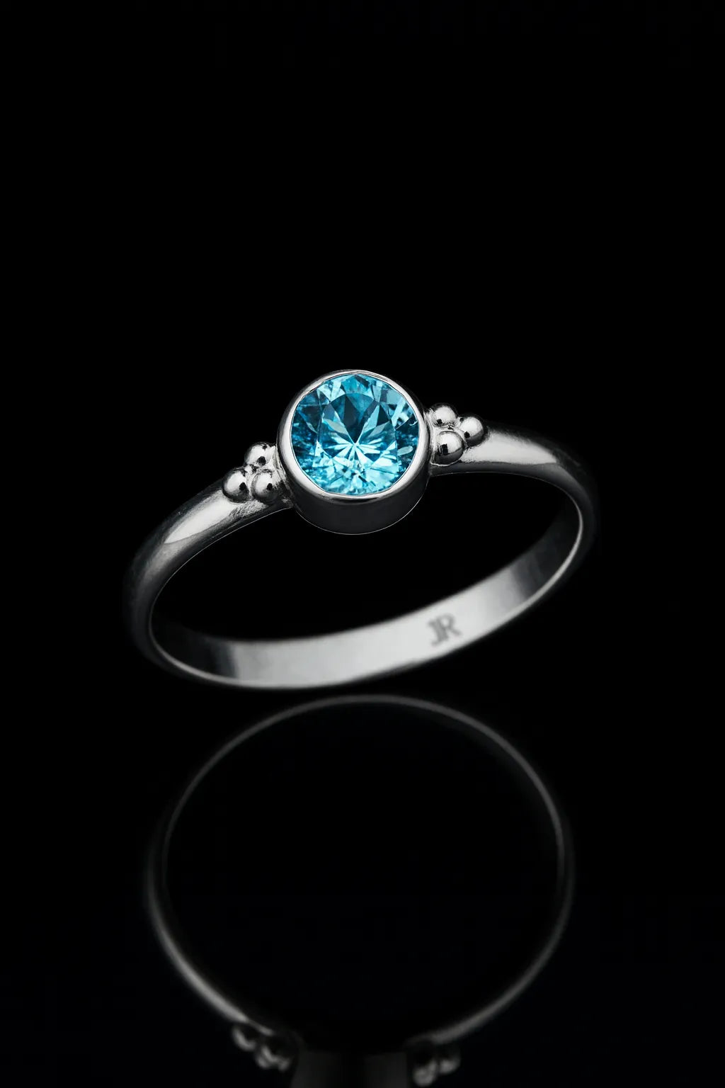 Silberring mit rundem blauem Topas in Zargenfassung, liegend auf schwarzem spiegelndem Untergrund mit klarer Reflexion, edles Schmuck Produktfoto in leicht schräger Perspektive.