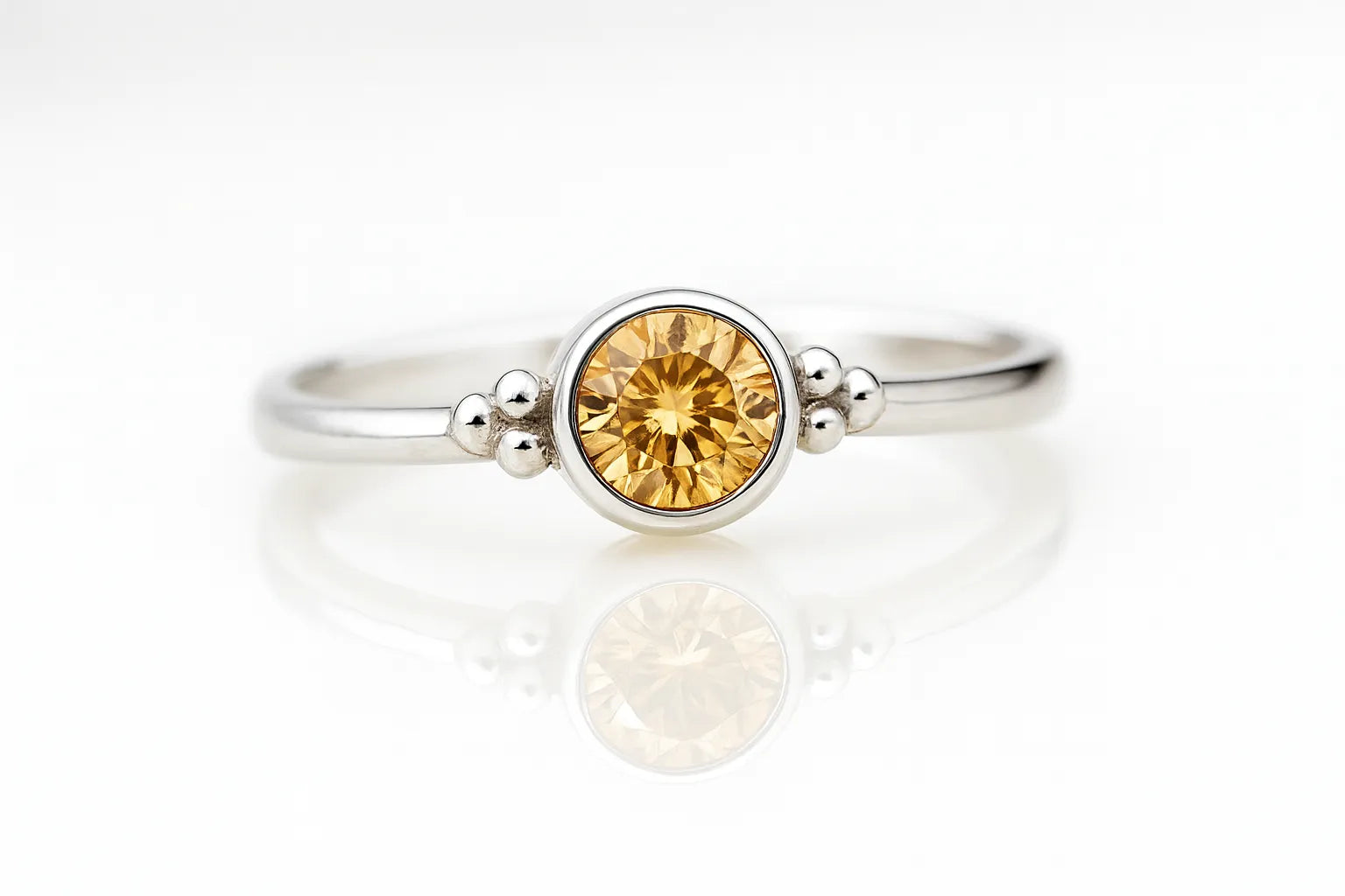 Damenring aus Sterlingsilber mit rund geschliffenem Citrin in Zargenfassung und dekorativen Silberkügelchen, liegend auf hellem spiegelnden Untergrund in seitlicher Produktansicht, edler Schmuck mit gelbem Edelstein.