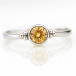 Damenring aus Sterlingsilber mit rund geschliffenem Citrin in Zargenfassung und dekorativen Silberkügelchen, liegend auf hellem spiegelnden Untergrund in seitlicher Produktansicht, edler Schmuck mit gelbem Edelstein.