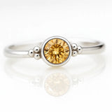 Damenring aus Sterlingsilber mit rund geschliffenem Citrin in Zargenfassung und dekorativen Silberkügelchen, liegend auf hellem spiegelnden Untergrund in seitlicher Produktansicht, edler Schmuck mit gelbem Edelstein.