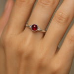 Nahaufnahme einer Frauenhand mit einem eleganten Silberring, der einen tiefroten, runden Granat-Cabochon als Mittelstein trägt. Der 4 mm große Edelstein ist in eine glatte Fassung eingebettet und reflektiert sanft das Licht. Der Fokus des Bildes liegt auf dem Ring, während der Hintergrund neutral und weich gehalten ist, um den Glanz des Silbers und die warme Farbe des Granats hervorzuheben.