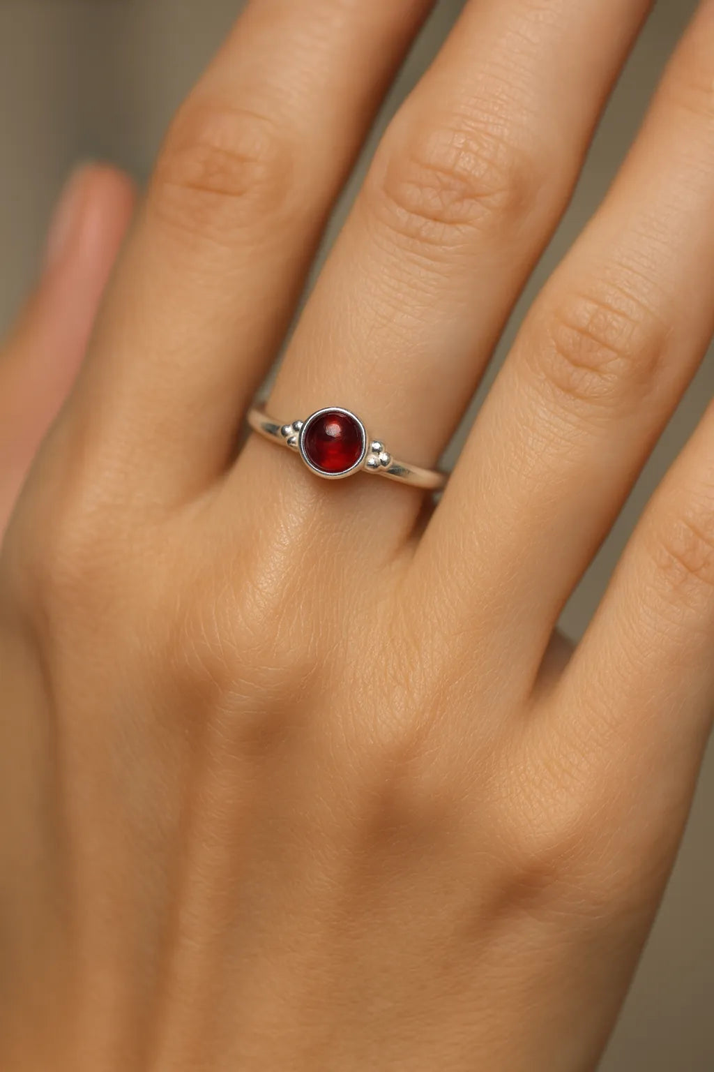 Nahaufnahme einer Frauenhand mit einem eleganten Silberring, der einen tiefroten, runden Granat-Cabochon als Mittelstein trägt. Der 4 mm große Edelstein ist in eine glatte Fassung eingebettet und reflektiert sanft das Licht. Der Fokus des Bildes liegt auf dem Ring, während der Hintergrund neutral und weich gehalten ist, um den Glanz des Silbers und die warme Farbe des Granats hervorzuheben.