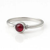 Feiner Damenring aus 925 Sterling Silber mit rundem Granat-Cabochon in tiefem Rot, eingefasst in einer Zargenfassung mit Silberkügelchen-Details, hochwertig fotografiert auf weißem spiegelnden Hintergrund als eleganter Edelsteinschmuck.