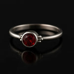 `Eleganter Damenring aus 925 Sterling Silber mit rund facettiertem Granat (4 mm) in Zargenfassung und seitlichen Silberkügelchen, edel inszeniert auf schwarzem spiegelnden Hintergrund mit weichem Studiolicht als hochwertiges Schmuckfoto.`