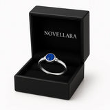 Ein silberner Ring mit einem runden, blauen 4-mm-Stein liegt in einer geöffneten schwarzen Schmuckschatulle mit weicher Samteinlage. Auf der Innenseite des Deckels steht das Logo „NOVELLARA“ in heller Schrift. Freigestellt vor hellem Hintergrund, fotografiert in Studioqualität.