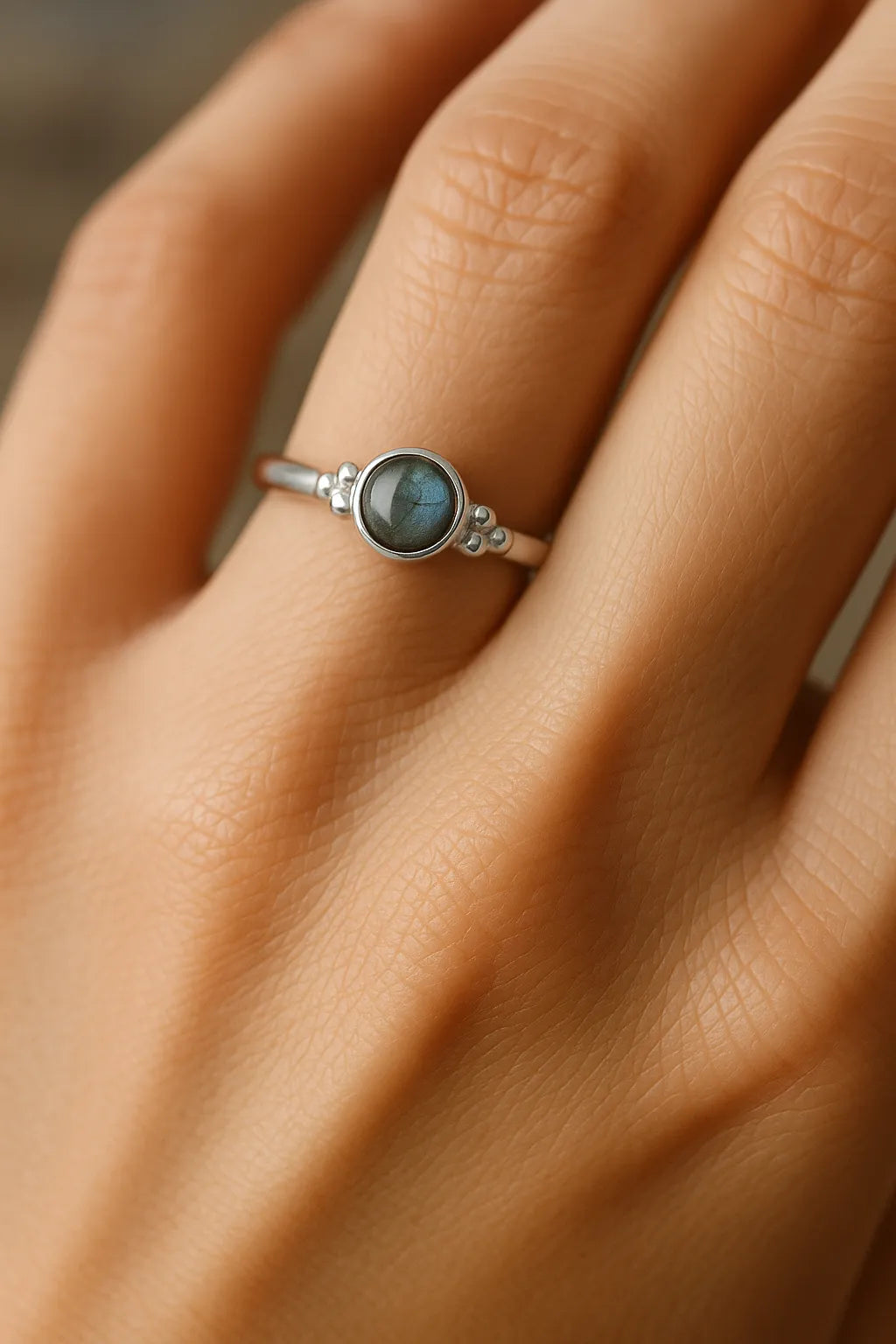 „Nahaufnahme einer Frauenhand, die einen eleganten Silberring mit einem runden Labradorit-Cabochon trägt. Der graublaue Stein zeigt ein sanftes Schimmern in Grün- und Blautönen und ist in eine feine, glänzende Ringschiene eingefasst. Der Fokus liegt auf dem Ring, der in natürlichem Licht dezent reflektiert.“