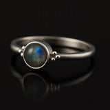 Damenring aus Sterling Silber mit rundem Labradorit Cabochon (4 mm) in Zargenfassung – filigraner Silberring mit blau-grün schimmerndem Edelstein, edel inszeniert auf schwarzem spiegelnden Hintergrund in seitlicher Perspektive.