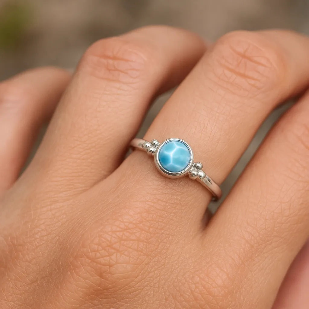 Eine Nahaufnahme einer Frauenhand zeigt einen silbernen Ring mit einem runden, hellblauen Larimar-Cabochon. Der 4 mm große Stein weist die charakteristische, wolkenartige Maserung des Larimars auf und ist in eine glatte Fassung mit kleinen silbernen Kugeldetails eingefasst. Weiches, natürliches Licht betont den Glanz des Steins und die polierte Oberfläche des Rings vor einem neutralen, unscharfen Hintergrund.