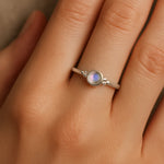 Nahaufnahme einer Frauenhand mit einem eleganten Silberring am Ringfinger. Der Ring zeigt einen etwa 4 mm großen, runden, facettierten Mondstein in einer glatten Zargenfassung, flankiert von kleinen Silberkügelchen. Der Mondstein schimmert mit einem zarten bläulichen Glanz, während das weiche Licht die natürliche Hautstruktur und den sanften Glanz des Silbers betont.