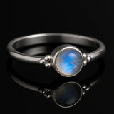 Edler Silberring mit schimmerndem blau-weißem Mondstein, liegend auf schwarzem spiegelndem Hintergrund mit starker Reflexion, hochwertiges Schmuck-Produktfoto seitlich aufgenommen.