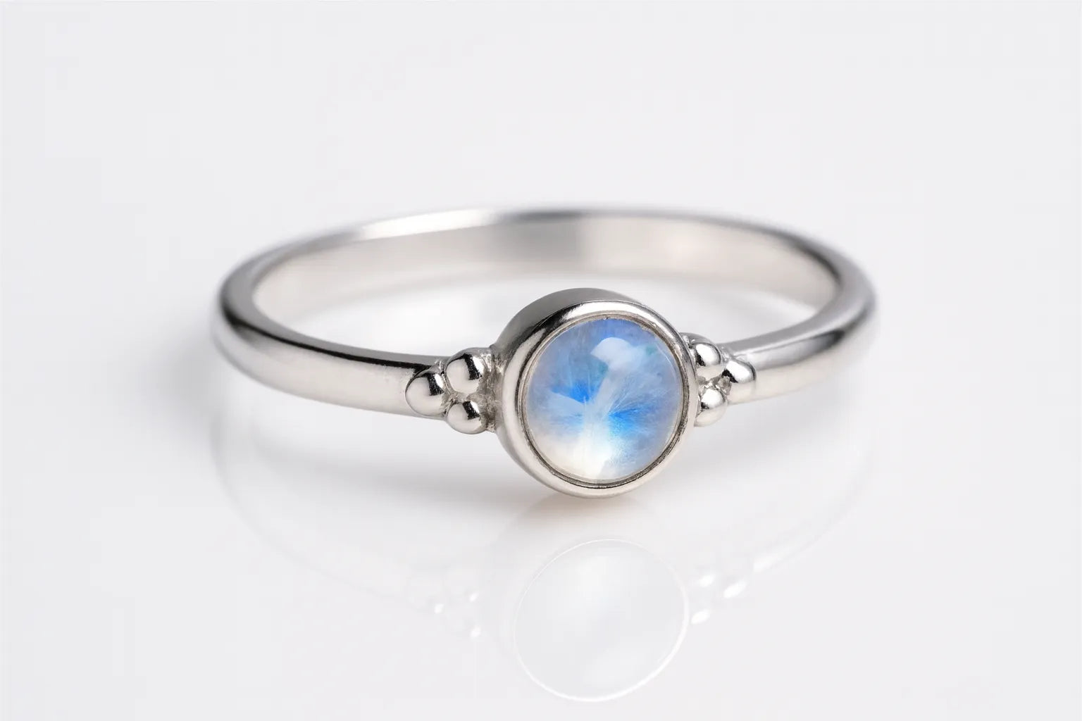 Zarter Silberring mit rundem Regenbogenmondstein in Zargenfassung, liegend auf hellem reflektierendem Untergrund, Produktfoto mit leichter seitlicher Perspektive für modernen Schmuck