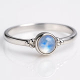Zarter Silberring mit rundem Regenbogenmondstein in Zargenfassung, liegend auf hellem reflektierendem Untergrund, Produktfoto mit leichter seitlicher Perspektive für modernen Schmuck
